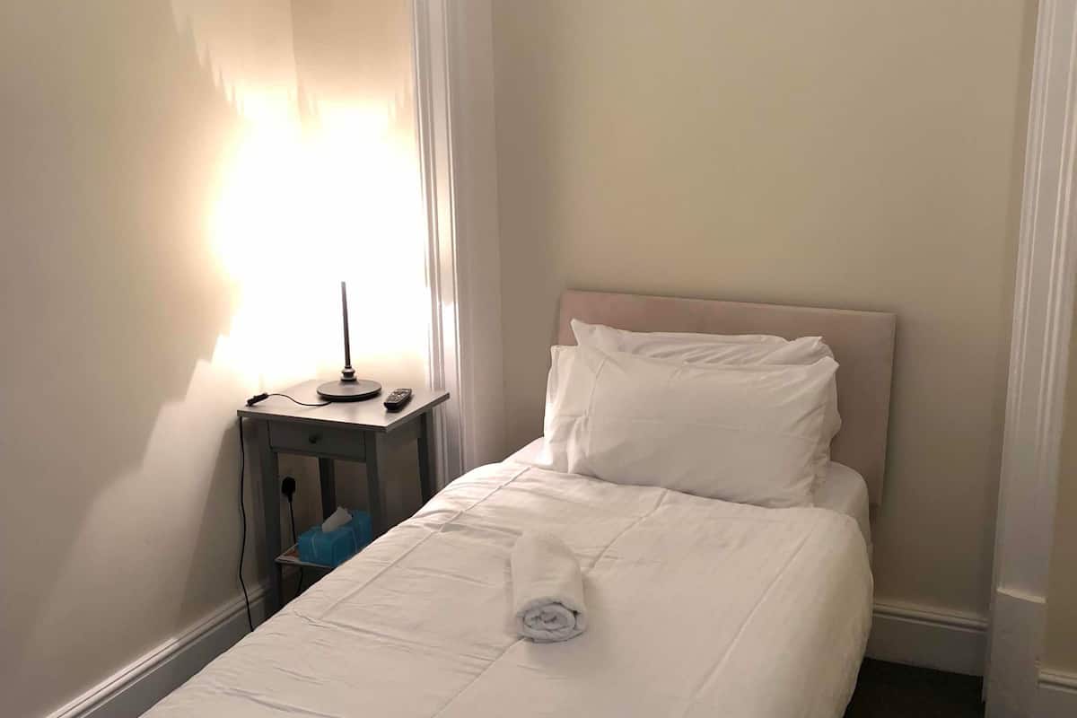 A neatly made bed with a white duvet and pillows, a bedside table with a lamp, and a wall-mounted shelf.