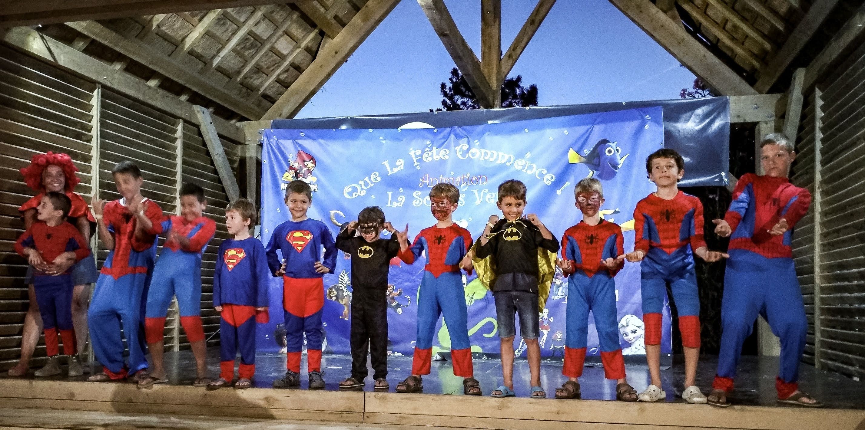 Un grupo de niños disfrazados de superhéroes de pie en fila sobre un escenario, con un fondo azul que muestra personajes de dibujos animados y texto.