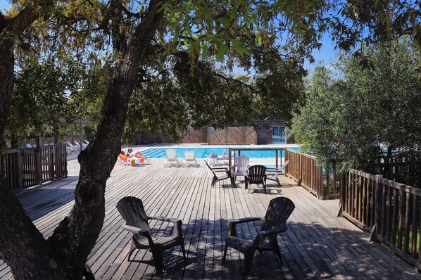 A wooden deck with lounge chairs, a pool, and a fenced area with a building in the background.
