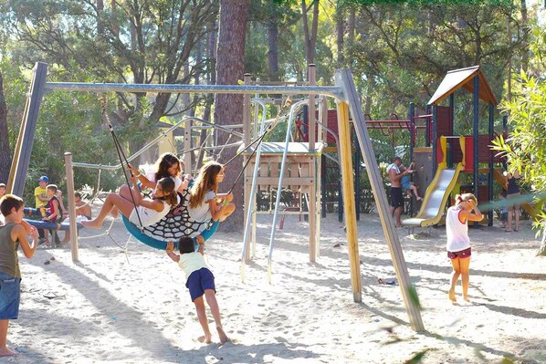 Un parque infantil con columpios y toboganes, rodeado de árboles y arena.