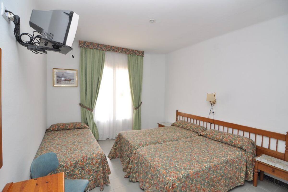 Una habitación con dos camas individuales, cabecera de madera, un escritorio, una silla, un televisor montado en la pared y una ventana con cortinas verdes.