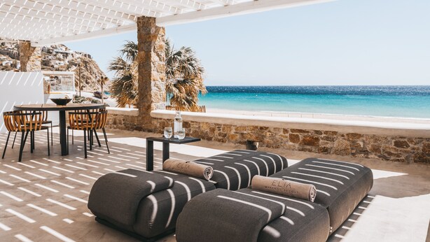 A beachside seating area with a stone wall, a table with chairs, and a view of the sea. - Elia Mykonos Resort (Mykonos)