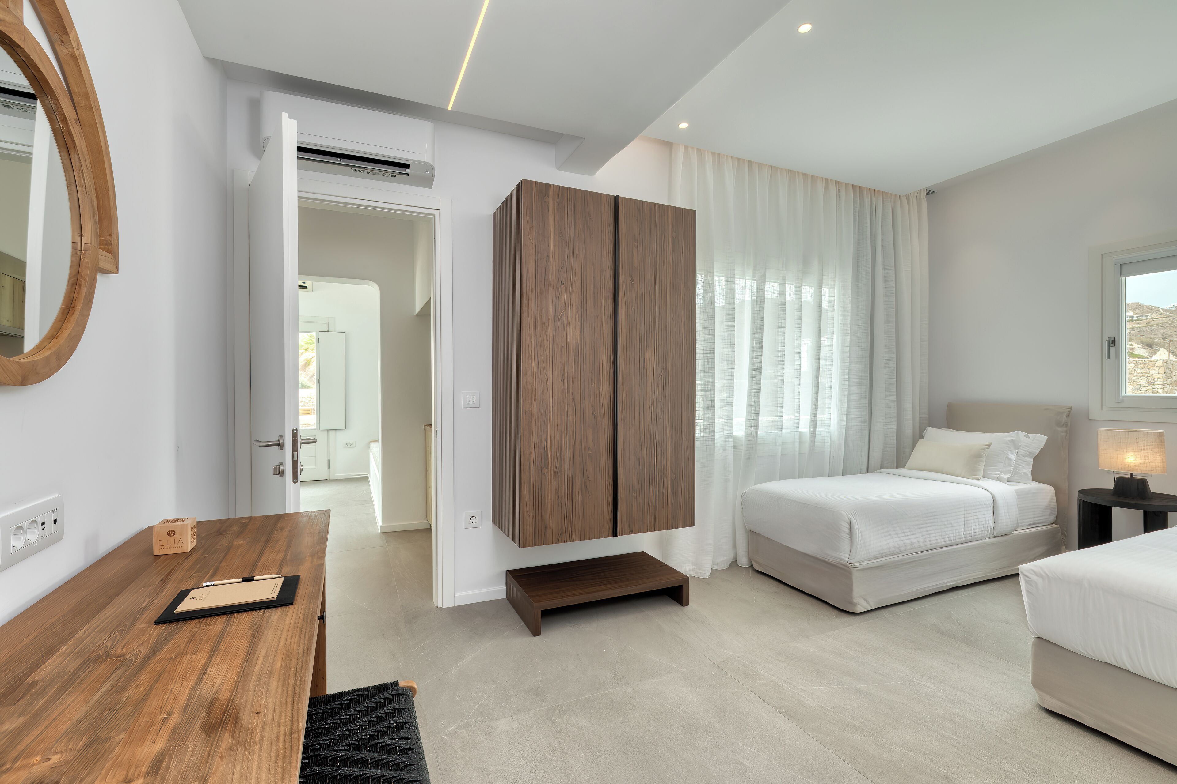 A modern bedroom with a wooden wardrobe, a bed, a desk, and a view of the outdoors.