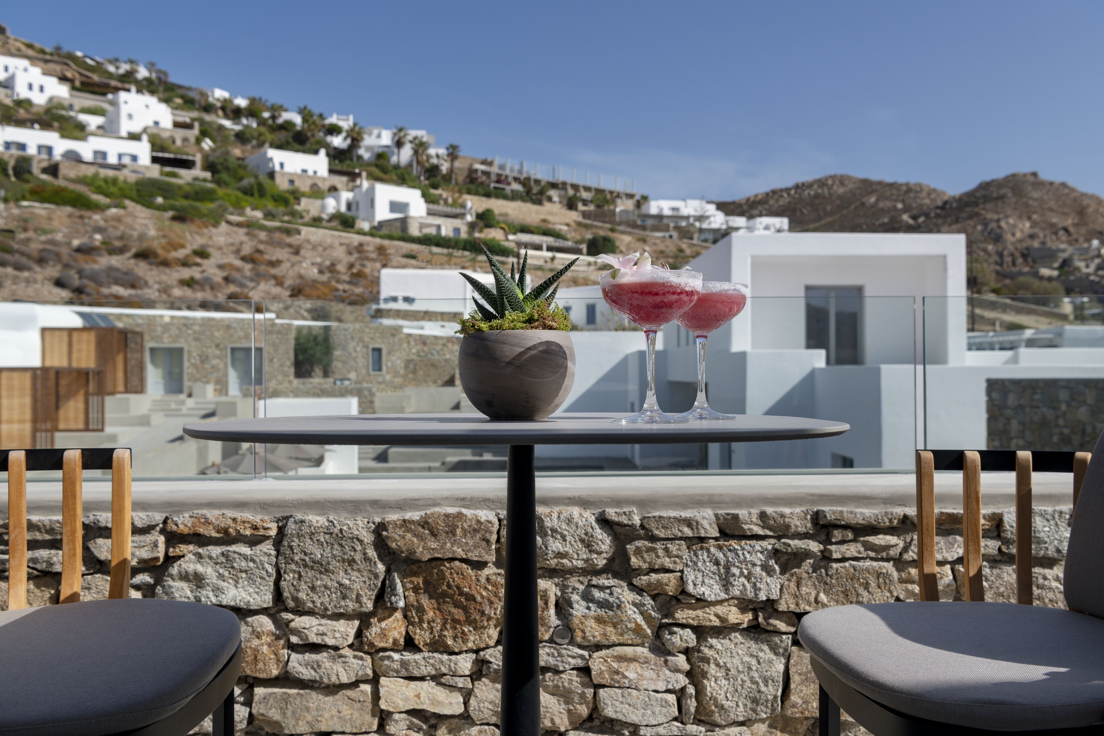Una terraza con pared de piedra, dos sillas, una mesa, una planta en maceta y dos cócteles.