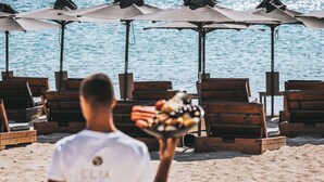 A beach with wooden lounge chairs and umbrellas, a person carrying a tray of food, and a clear view of the sea. - Elia Mykonos Resort (Mykonos)