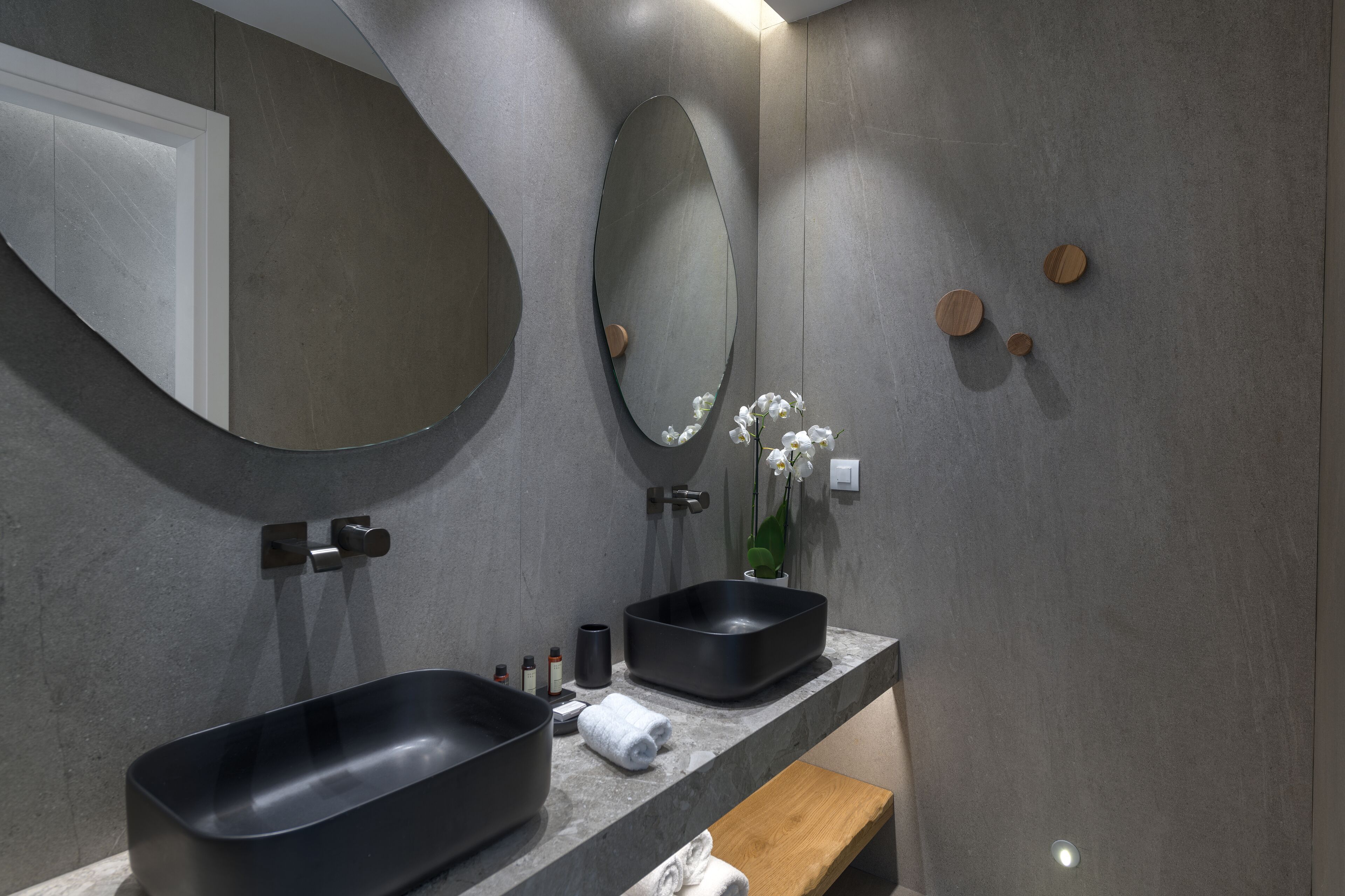 A modern bathroom with a large oval mirror, two black sinks, a wooden bench, and a vase with white flowers.
