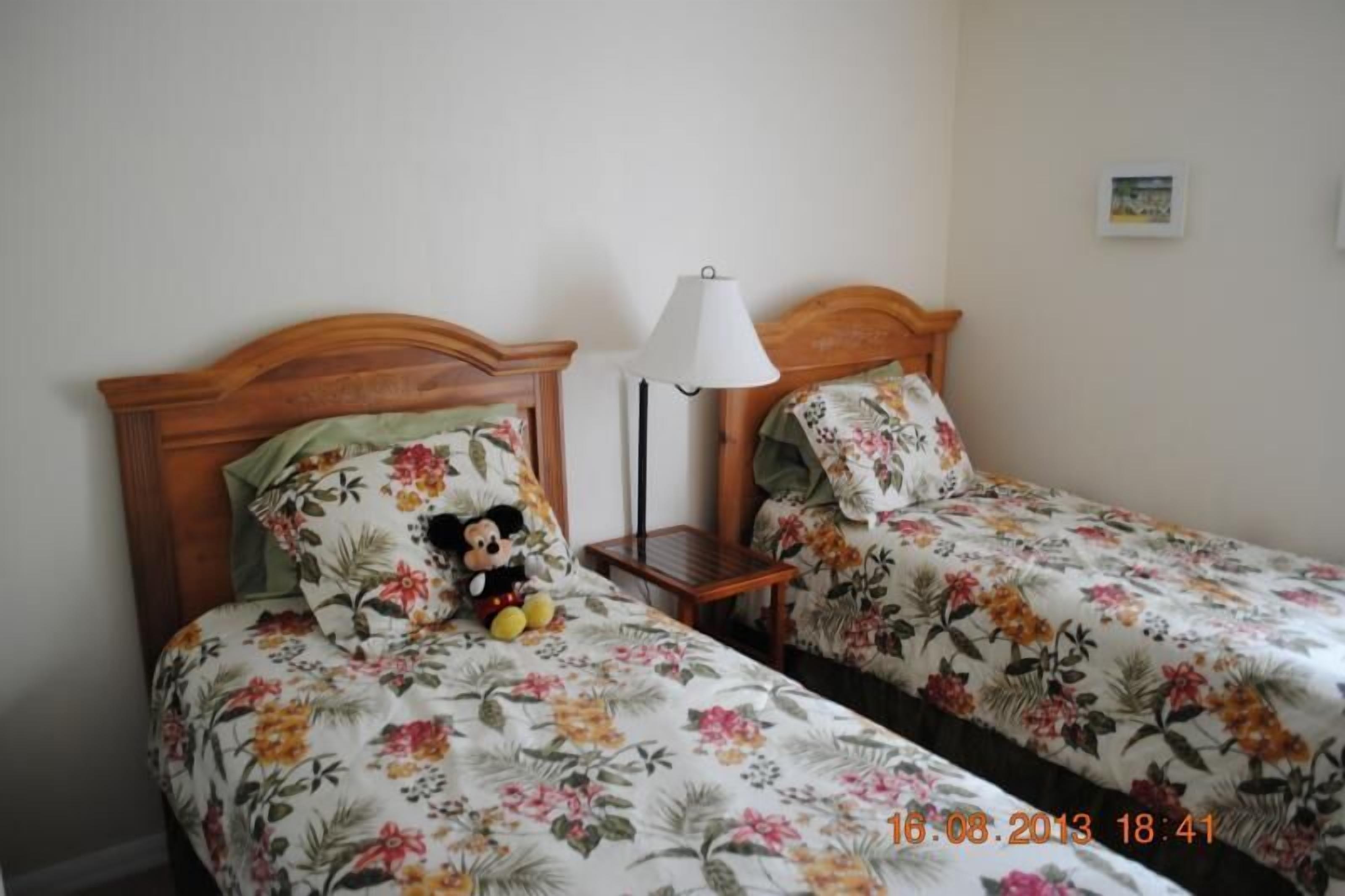 A room with two beds, each with floral bedding and a Mickey Mouse plush toy on one bed. There is a nightstand with a lamp between the beds.