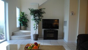 Een woonkamer met een televisie, een plant en een schaal fruit op een tafel.
