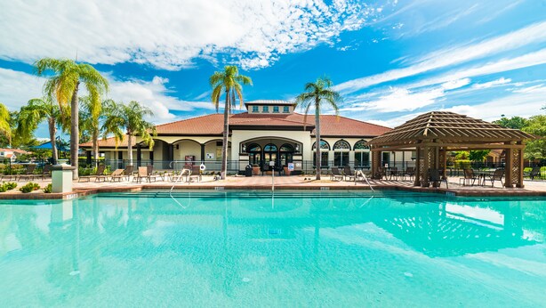 A swimming pool with lounge chairs and palm trees, a gazebo, and a building with a tiled roof.
