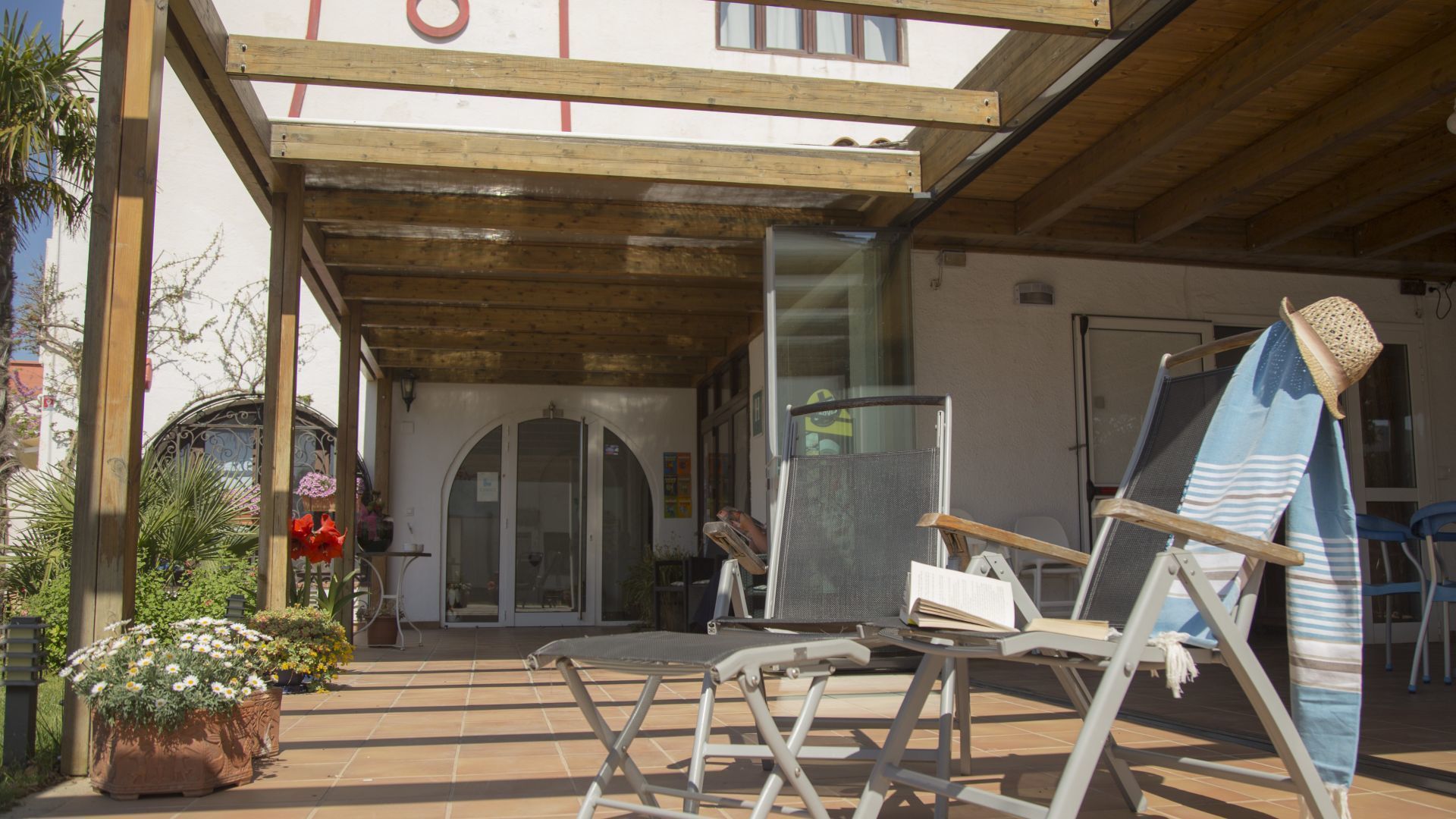 A covered outdoor patio with a wooden ceiling, a glass door entrance, and a seating area with a chair and a table.