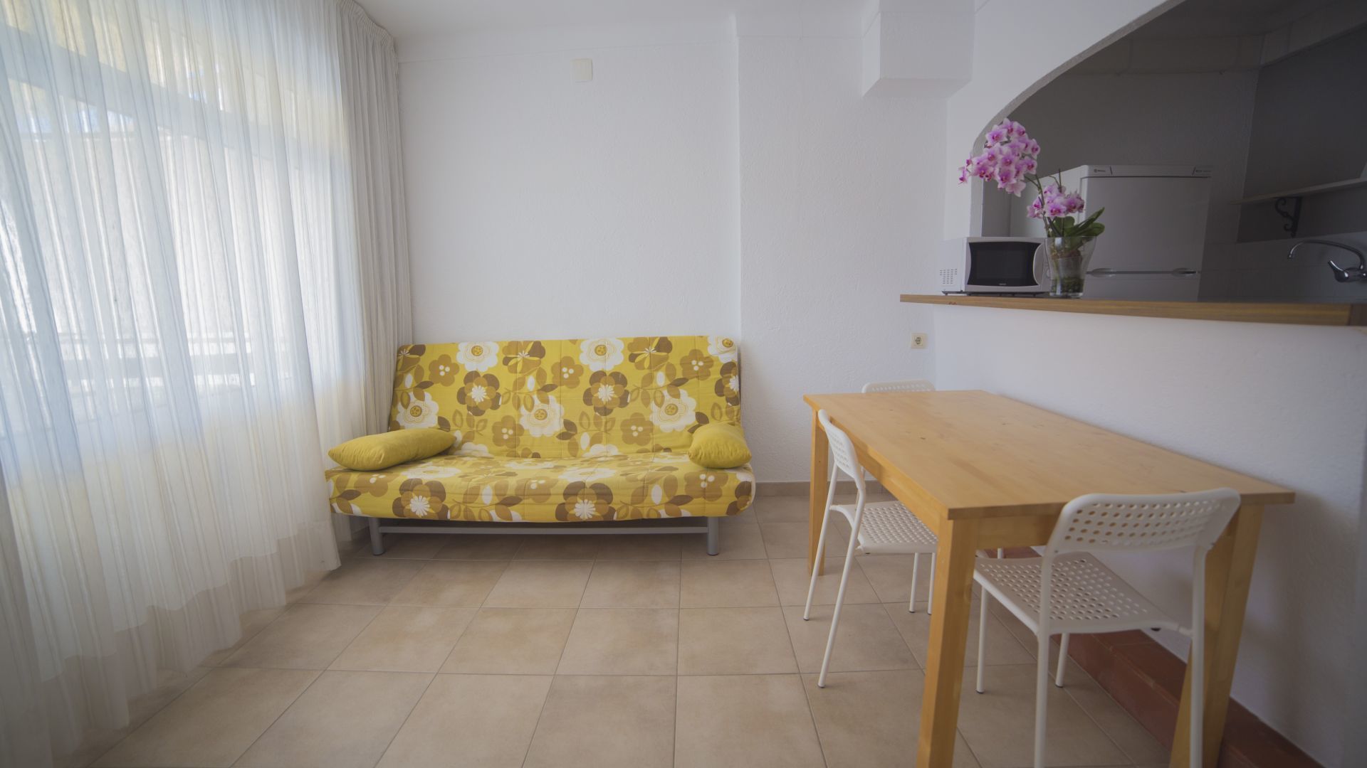 A small, bright room with a yellow floral sofa, a wooden table, and white chairs.