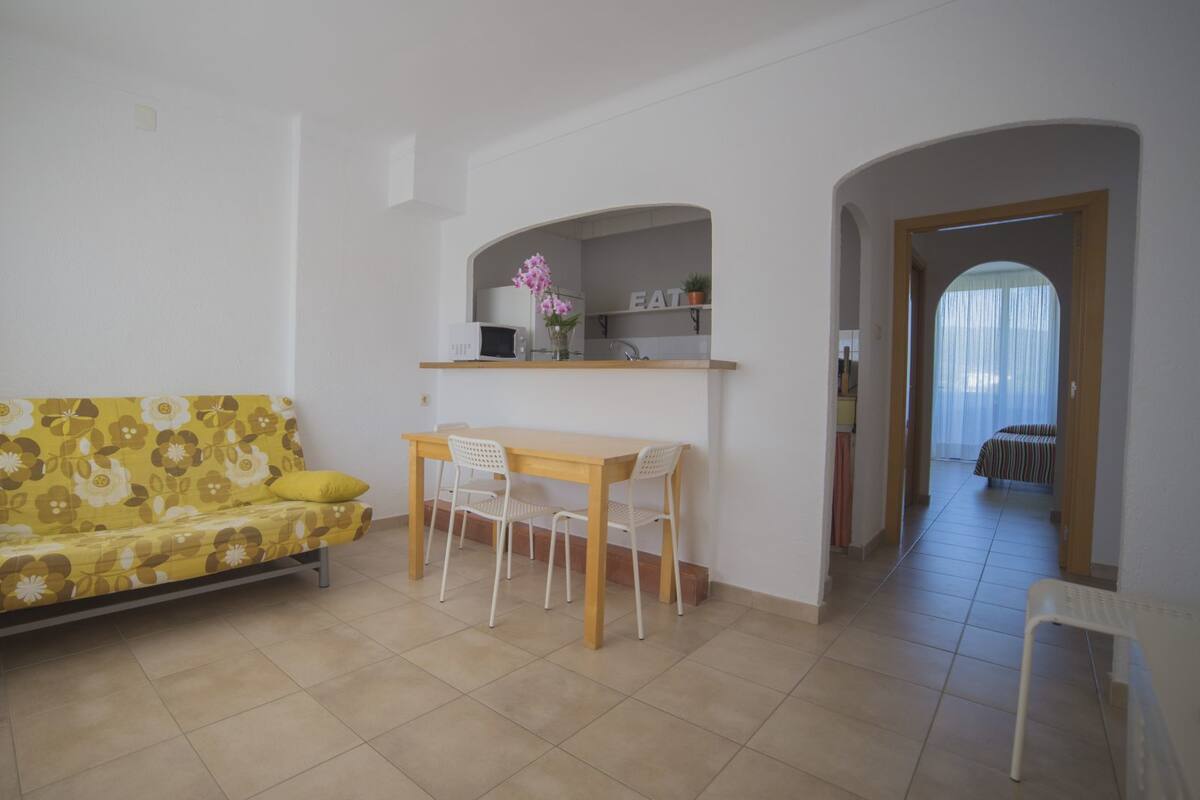 A living room with a sofa, a dining table with chairs, a kitchen area with a microwave, and a view of a balcony.