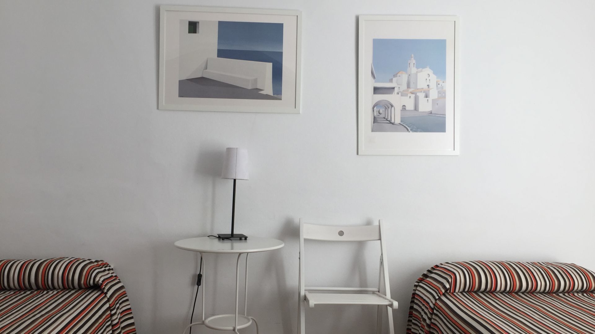 A room with two striped beds, a white chair, a small round table with a lamp, and two framed pictures on the wall.