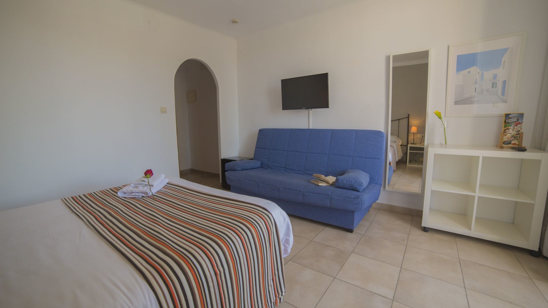 A hotel room with a bed, a blue sofa, a TV, and a small white shelf with a picture frame and a vase.