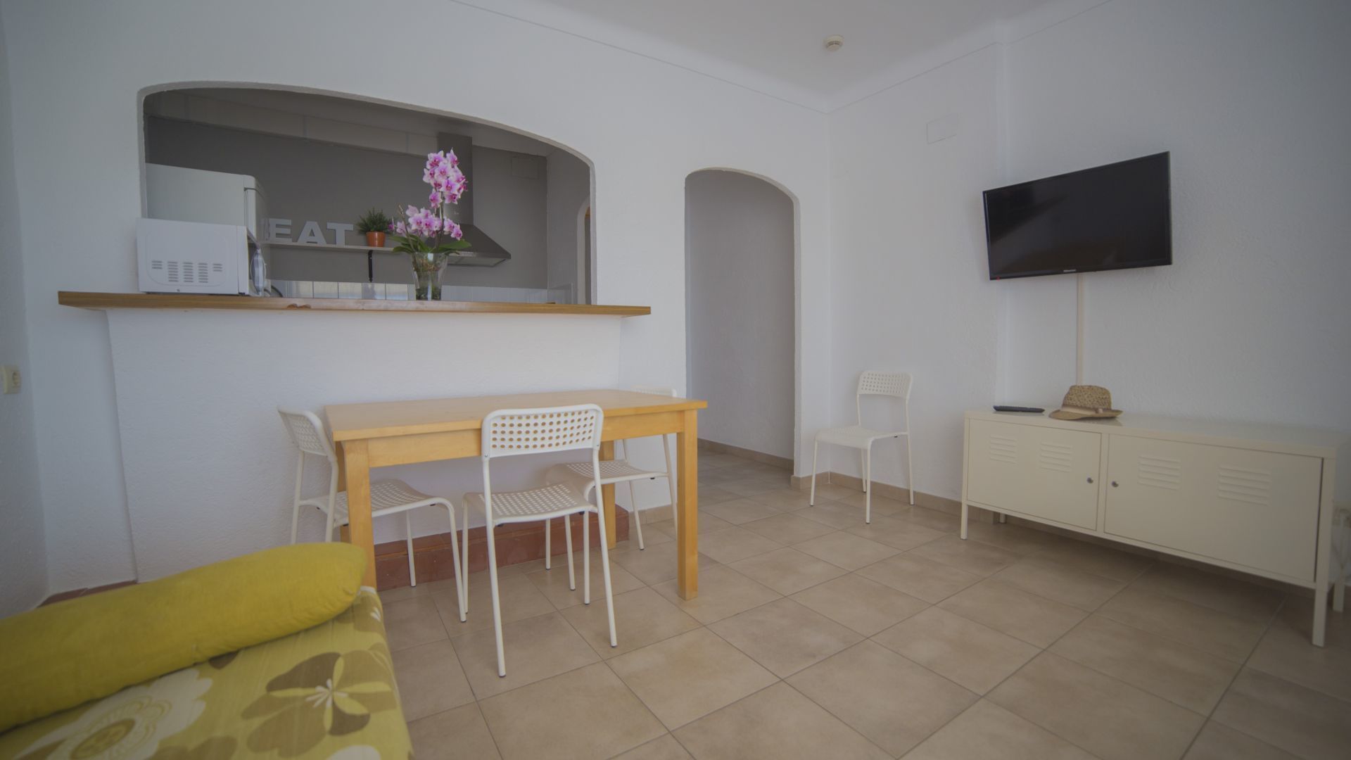 A small, bright room with a dining table, chairs, a sofa, a TV, and a kitchenette.
