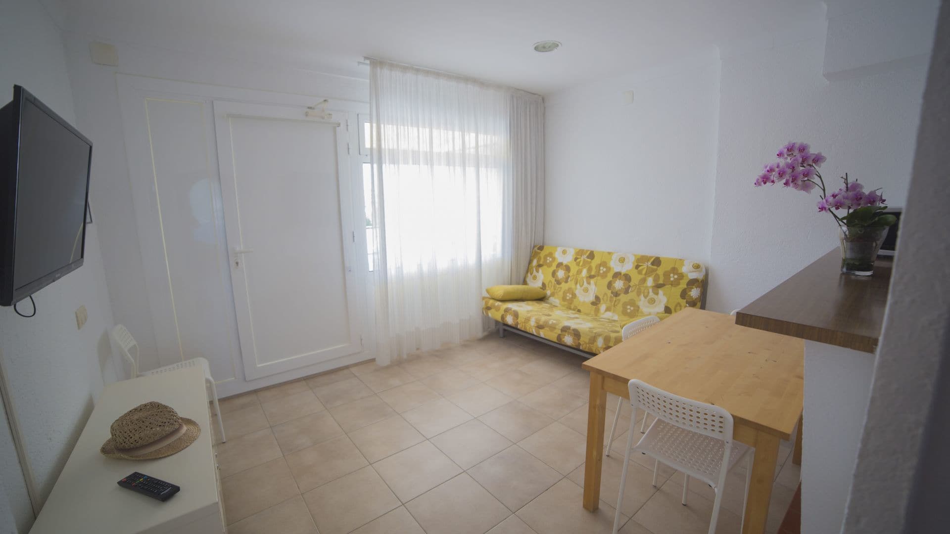 A small, bright room with a sofa, a table, a TV, and a window with curtains.