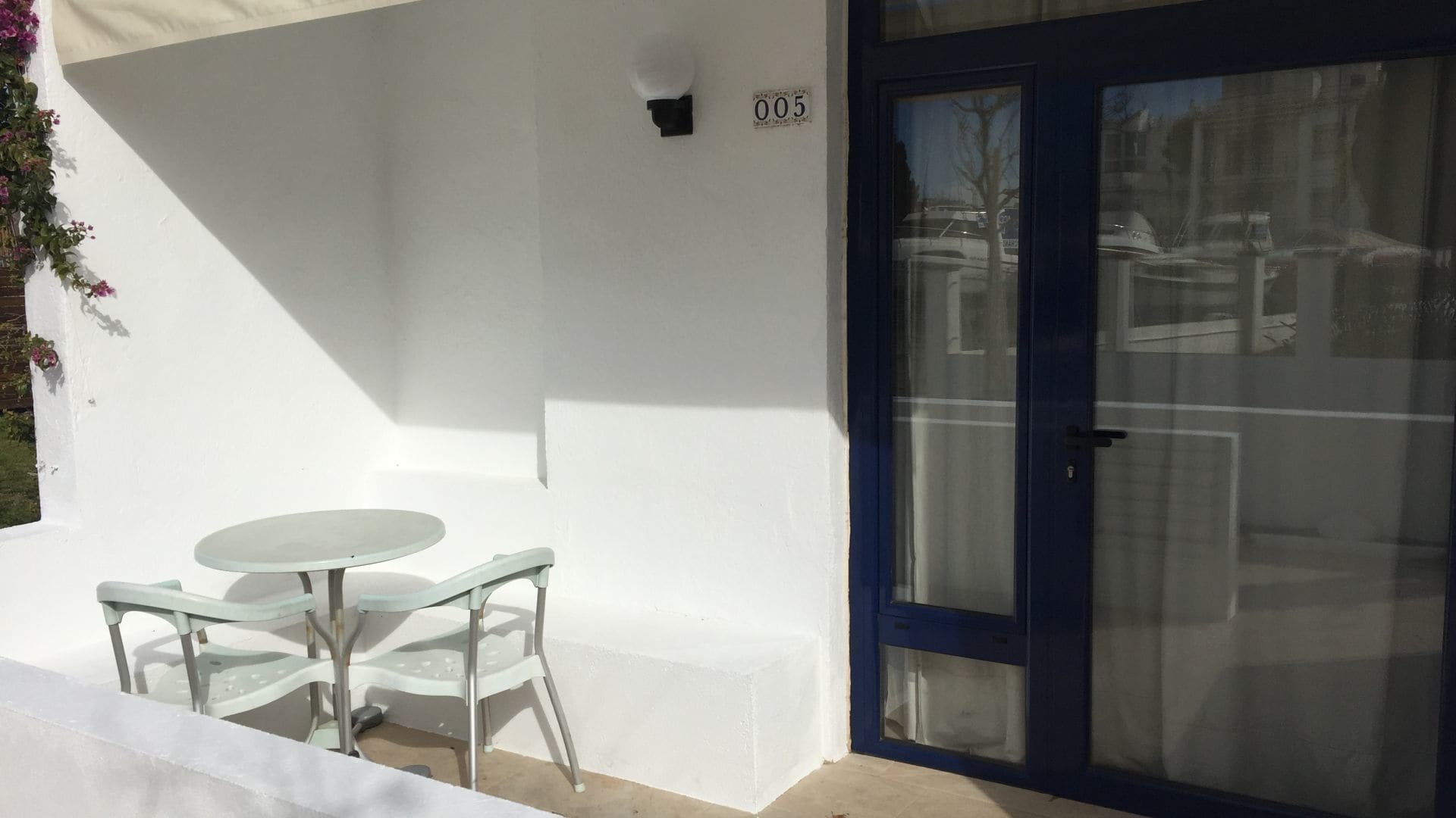A white exterior wall with a number '005' sign, a glass door, and a small round table with two chairs.