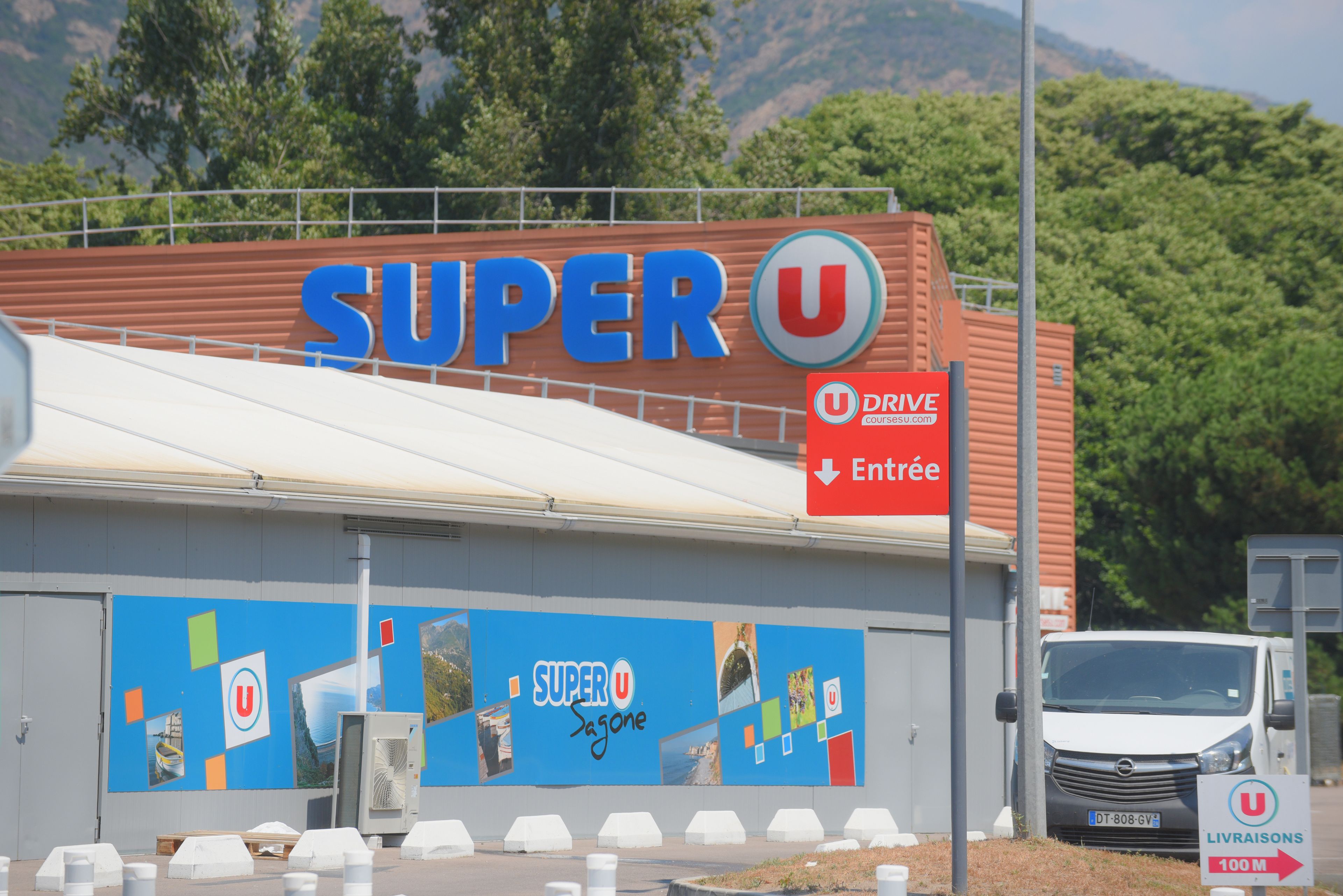 Un supermercado Super U con un letrero azul grande, un cartel de entrada para vehículos y un anuncio de entrega a domicilio.