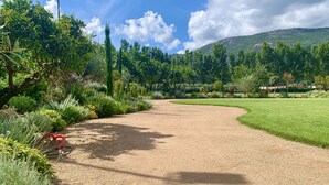 A gravel path in a landscaped garden with various plants and trees, leading towards a green field and a hill in the background. - Camping Le Sagone (Vico)