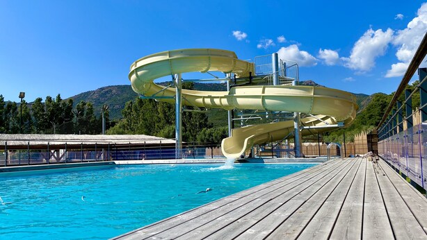 A water slide structure over a pool with a wooden deck and mountains in the background. - Camping Le Sagone (Vico)