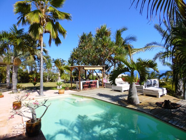 A poolside area with a wooden deck, lounge chairs, a barbecue setup, and palm trees. - La Kaz Nana Sakifo - Adults Only (Saint-Paul)