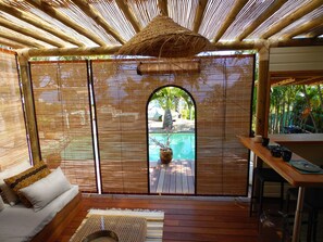 A room with a wooden floor, a sofa, a dining table, and a view of a pool through a bamboo screen. - La Kaz Nana Sakifo - Adults Only (Saint-Paul)