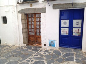A white building with a stone-paved entrance, a wooden door with glass panels, and a blue door with decorative paper cutouts. - Hostal Vehí (Cadaqués)