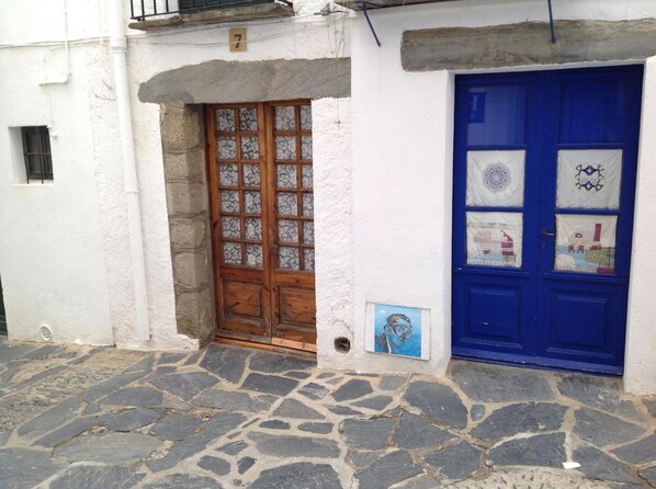 Un bâtiment blanc avec une entrée pavée de pierres, une porte en bois dotée de panneaux de verre et une porte bleue ornée de motifs découpés dans du papier.