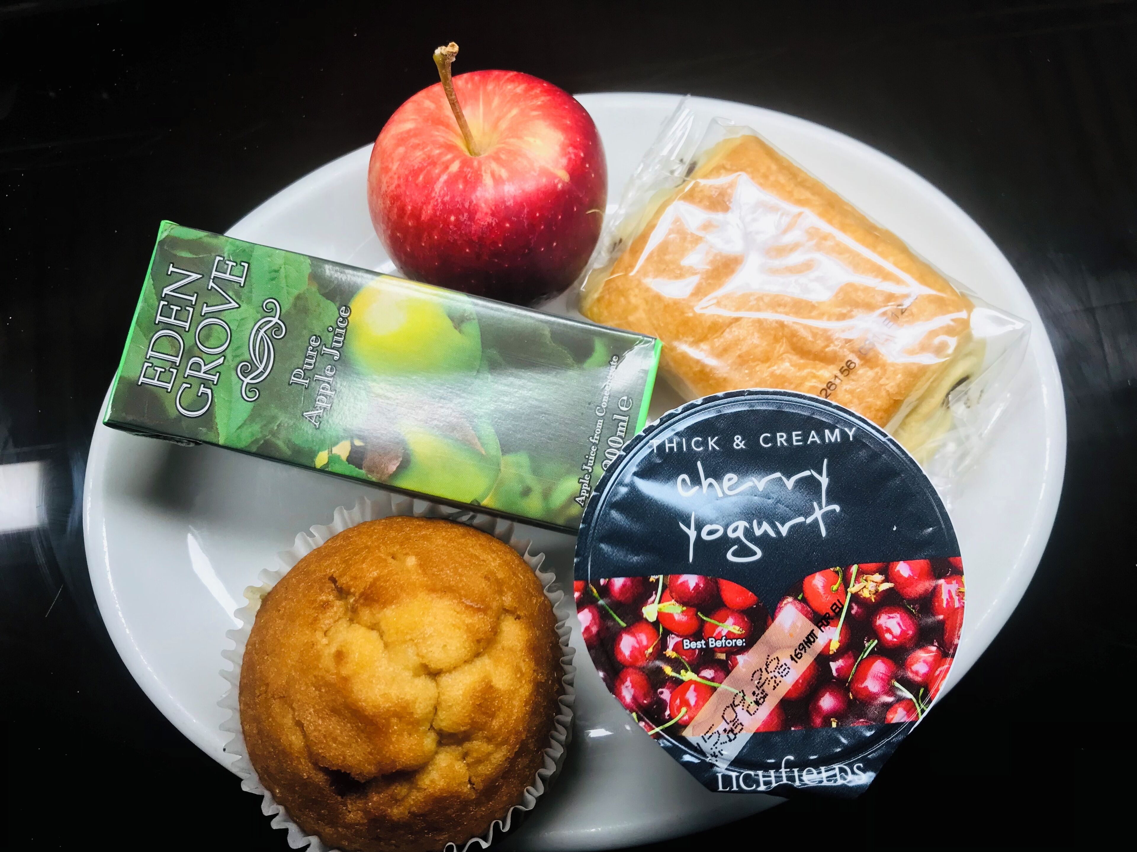 Lautasella on omena, muffinssi, voileipä, mehupakkaus ja jogurttipurkki.