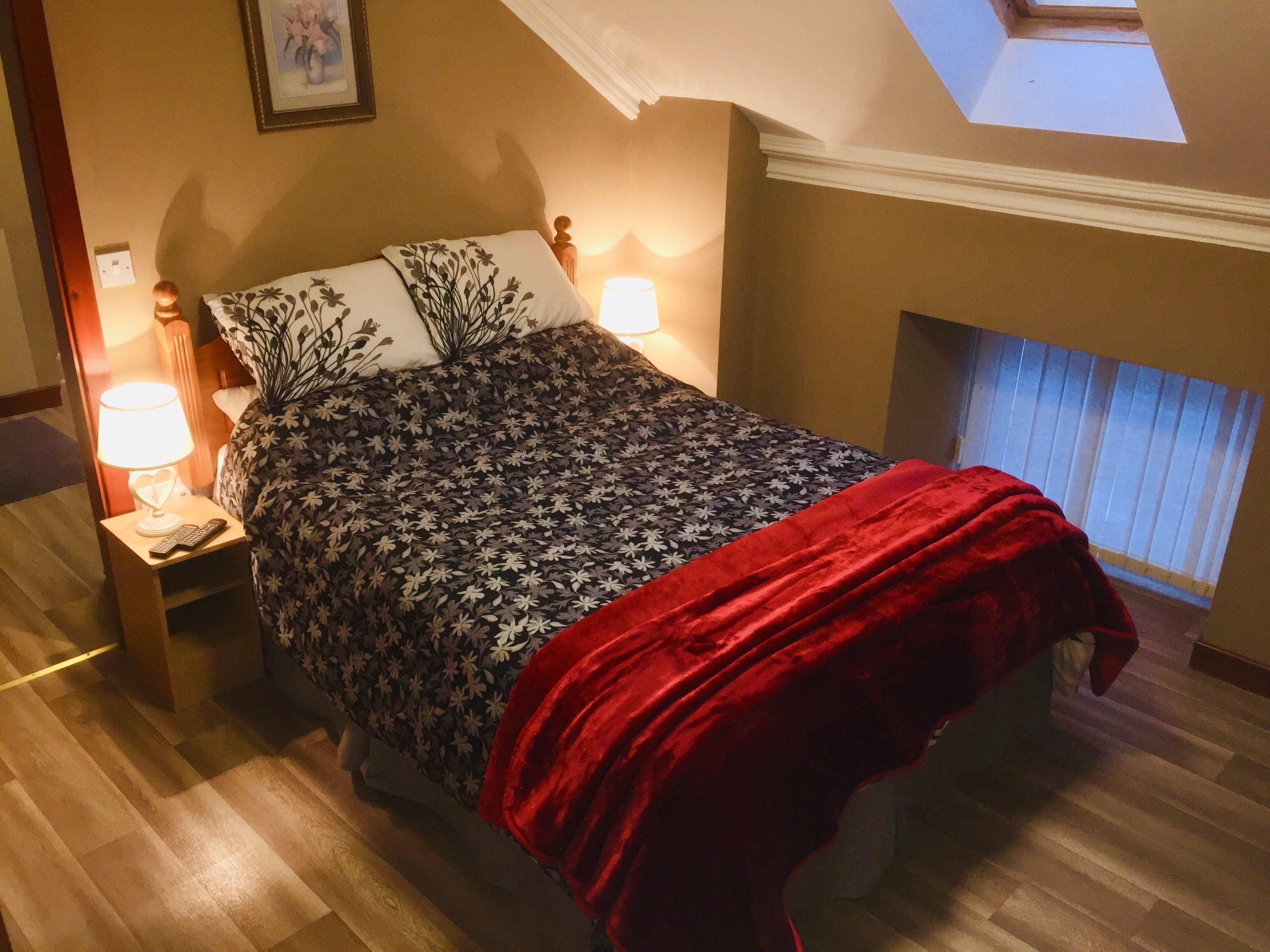 Ein Schlafzimmer mit einem Bett, zwei Nachttischlampen, einer roten Tagesdecke, einem Nachttisch, einem Wandbild und einem Oberlicht.