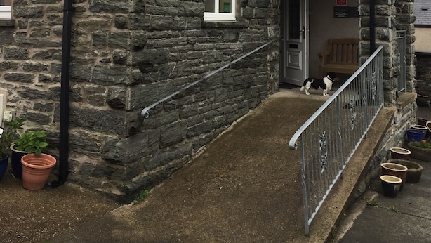 A stone building with a large arched window, a small porch with a bench, and a dog outside. - Pisgah Guest House Snowdonia (Blaenau Ffestiniog)