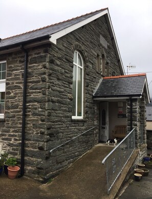 A stone building with a large arched window, a small porch with a bench, and a dog outside. - Pisgah Guest House Snowdonia (Blaenau Ffestiniog)
