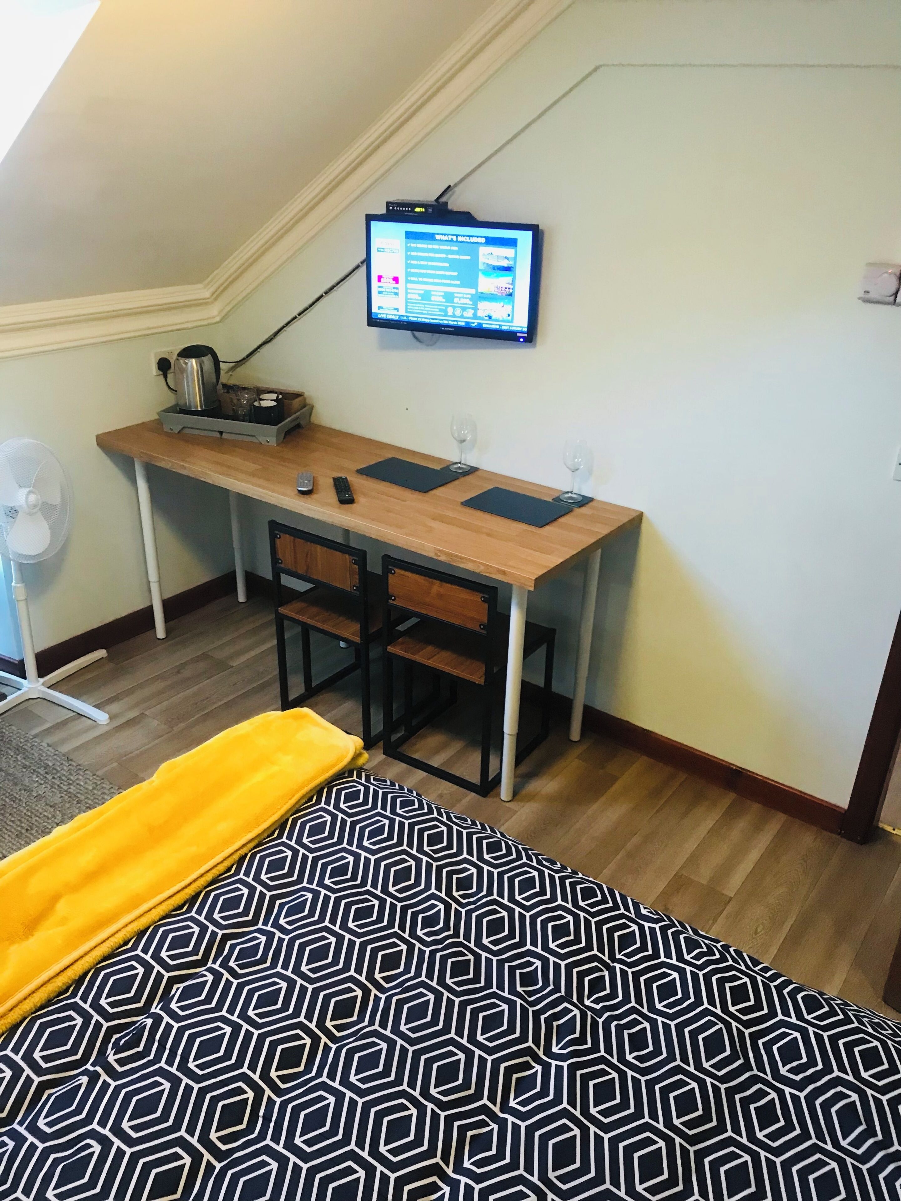 A room with a desk, chairs, a TV, a fan, and a bed with a patterned bedspread.