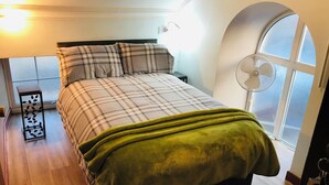 A bedroom with a bed, a bedside table, a window with a view, and a standing fan. - Pisgah Guest House Snowdonia (Blaenau Ffestiniog)