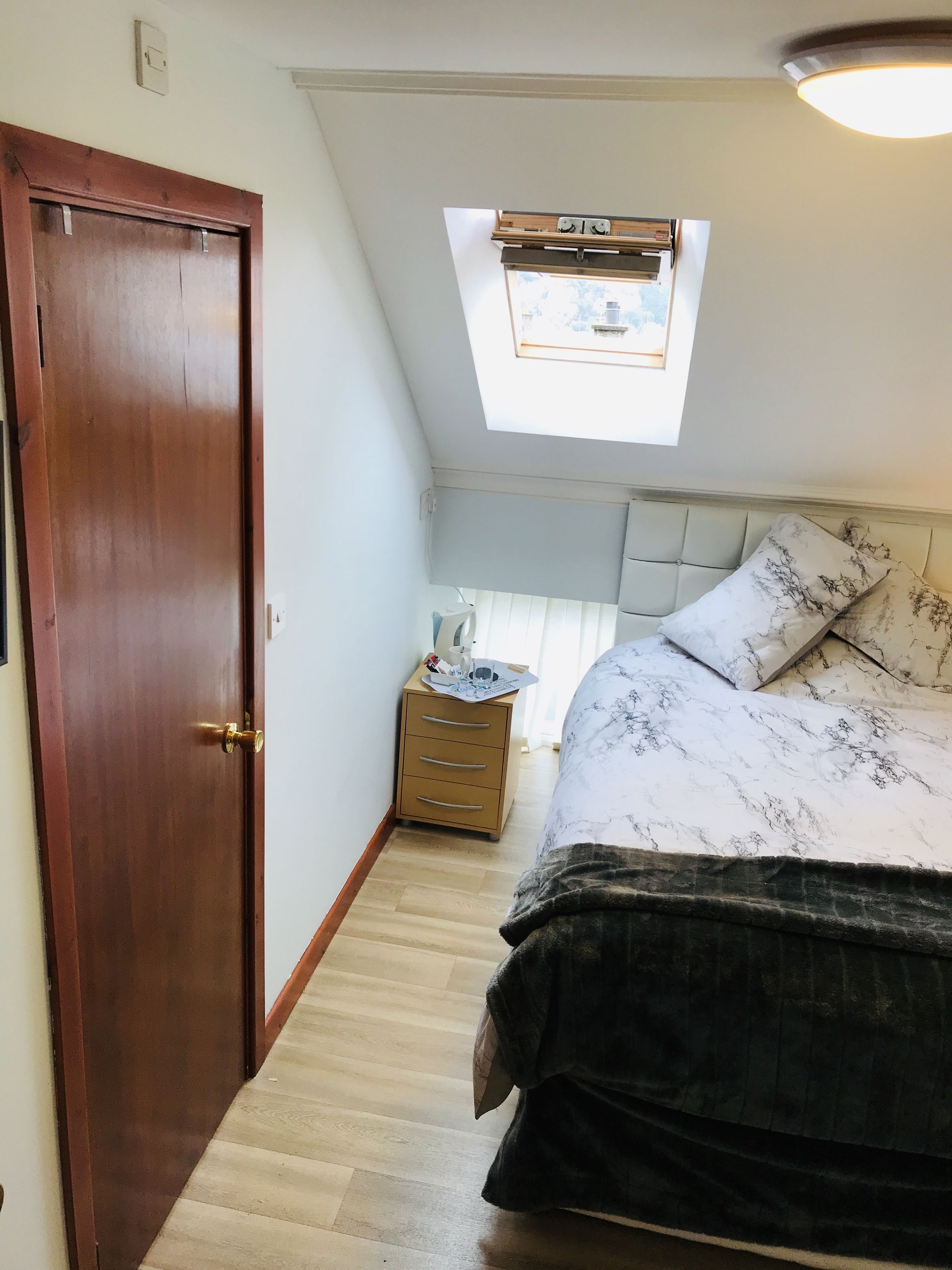 A bedroom with a wooden door, a bedside table with a lamp, a bed with a patterned bedspread, and a skylight above the bed.