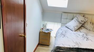 A bedroom with a wooden door, a bedside table with a lamp, a bed with a patterned bedspread, and a skylight above the bed.