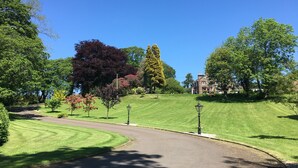 A well-maintained lawn with a curved driveway, surrounded by mature trees and a large building in the background. - Fludha Guest House (Kirkcudbright)