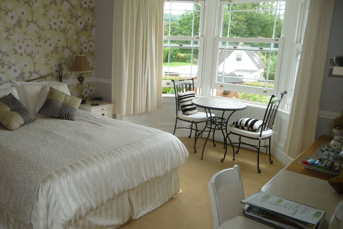 A bedroom with a large bed, a bedside table with a lamp, a window with curtains, a small round table with chairs, and a view of the outside.