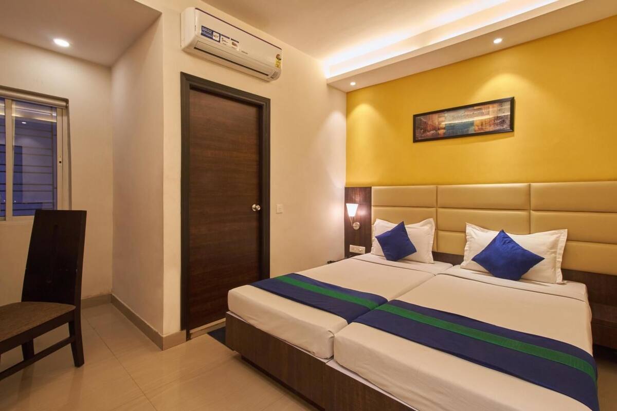A hotel room with a double bed, a nightstand, a chair, an air conditioning unit, a door, and a wall-mounted picture.