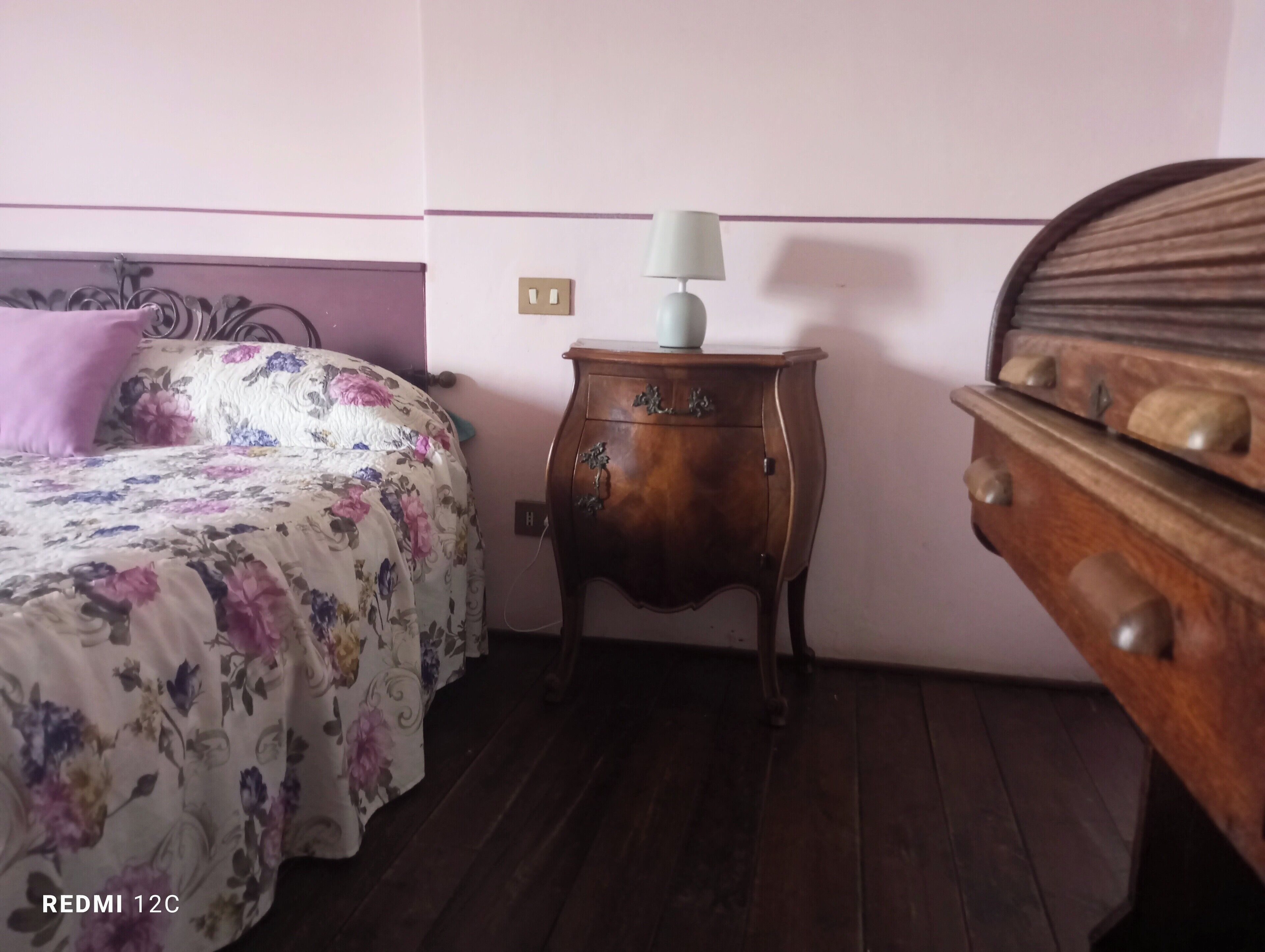 Et soveværelse med et blomstret sengetæppe, et trænatbord med en lampe og et kommode.