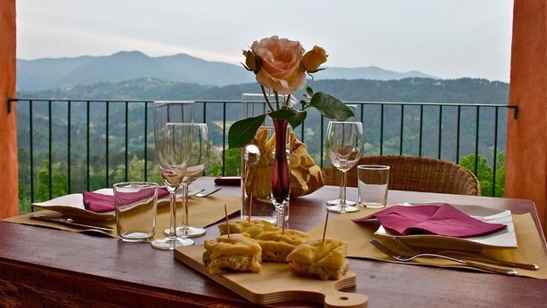 Mesa preparada para dos con un florero con una sola rosa, dos copas de vino y un plato de comida, con vistas a un paisaje montañoso.