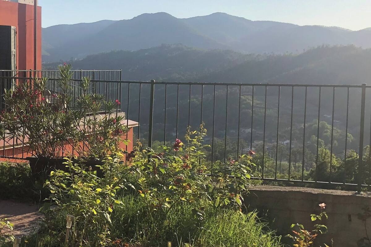 A balcony with a view of mountains, a red building with shutters, and outdoor furniture.