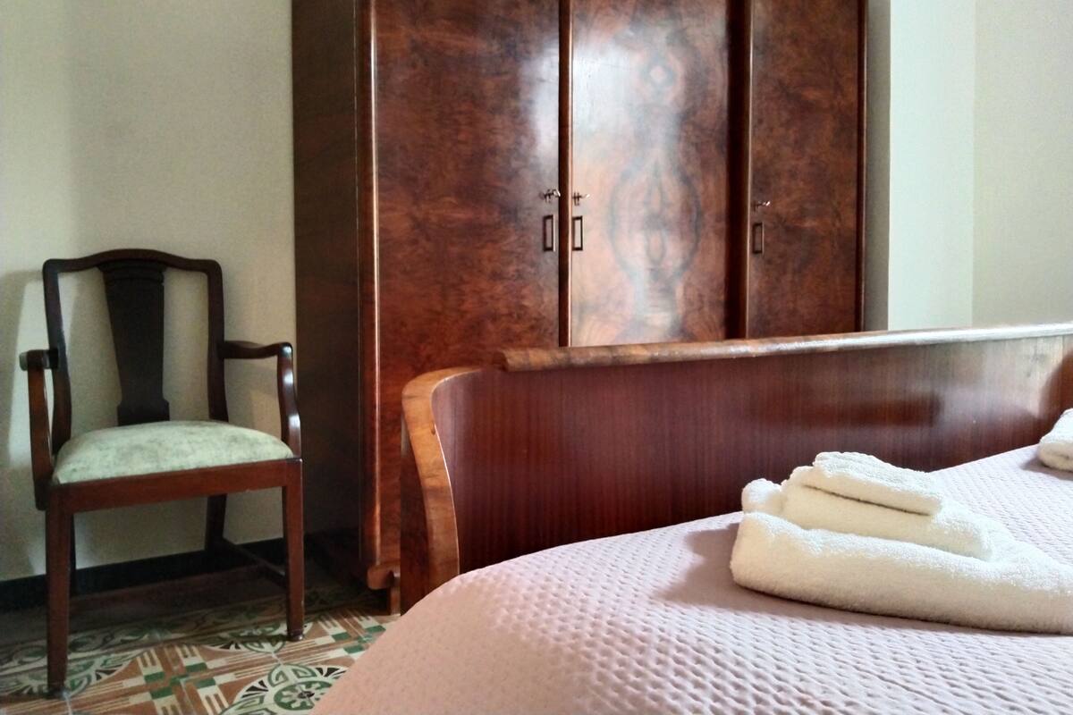 A hotel room with a wooden wardrobe, a wooden chair with a cushioned seat, and a neatly made bed with folded towels.