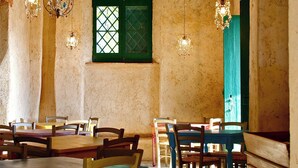 Un comedor con mesas y sillas de madera, un ventanal verde con diseño de celosía y lámparas colgantes.