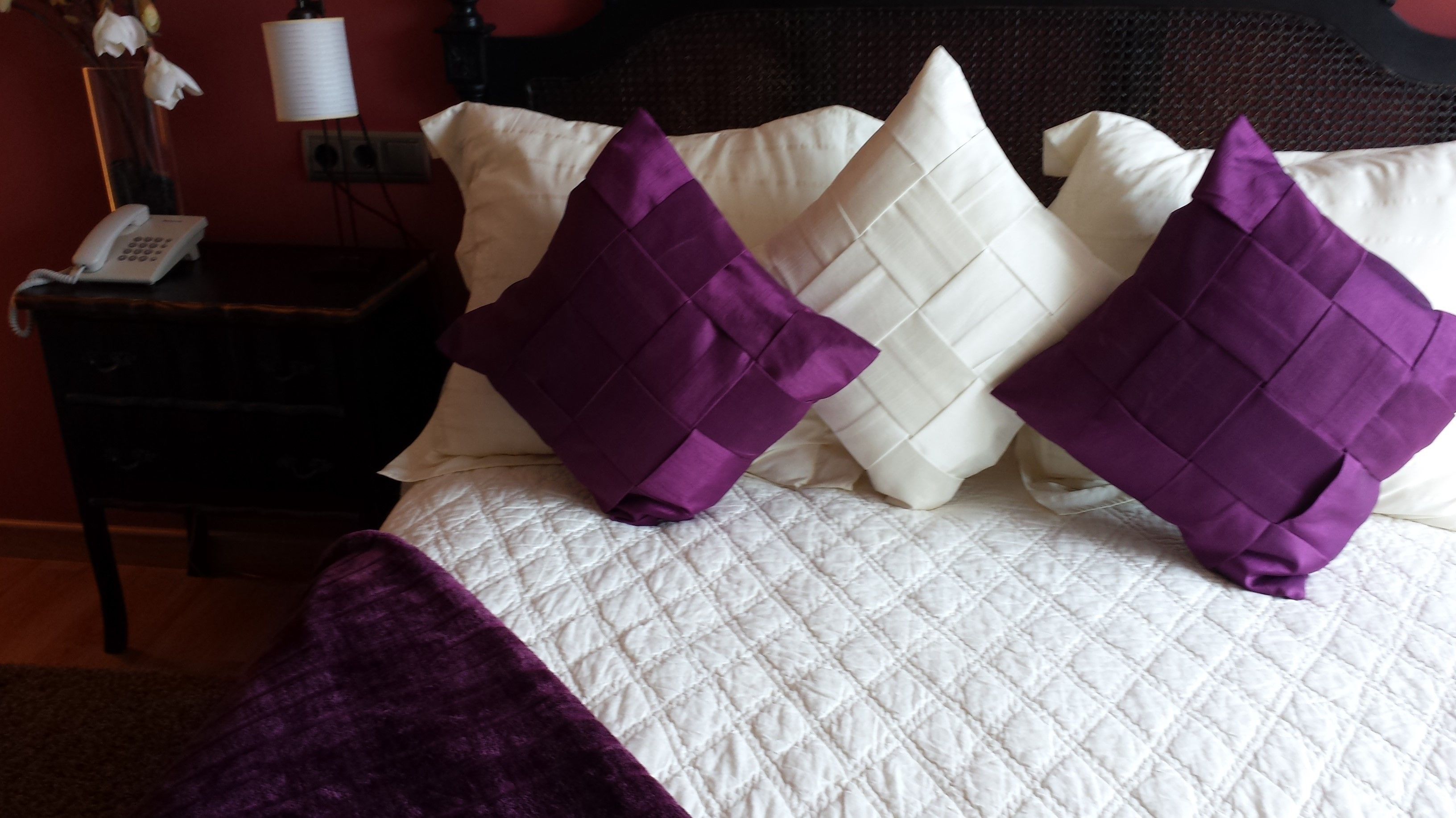 A bed with a white quilted bedspread, two purple pillows, and a dark wooden nightstand with a white telephone.