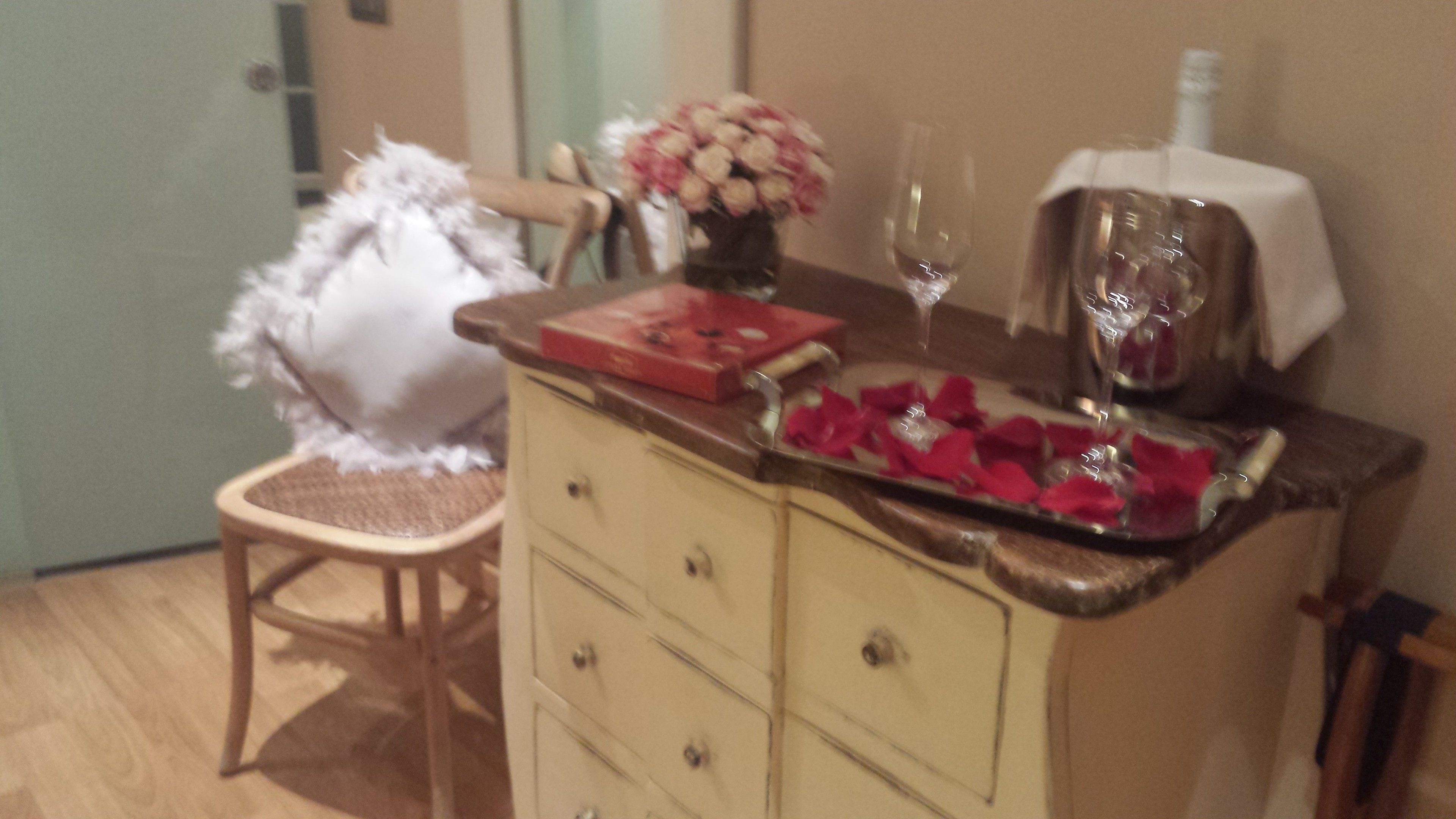 A small room with a wooden chair, a dresser with drawers, a vase with flowers, a tray with rose petals, and a bottle on the dresser.