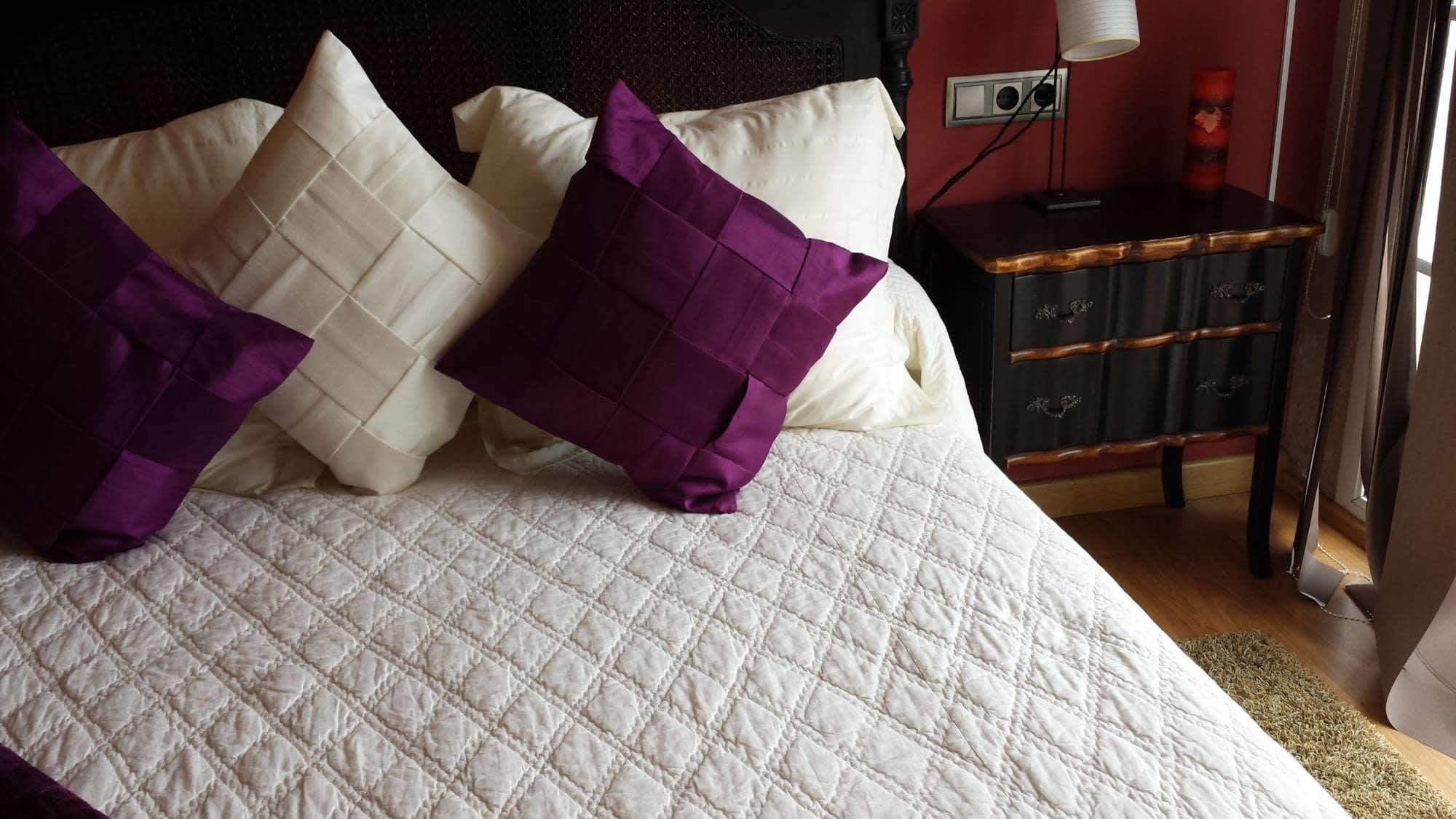 A bed with a white quilted bedspread and two purple pillows.