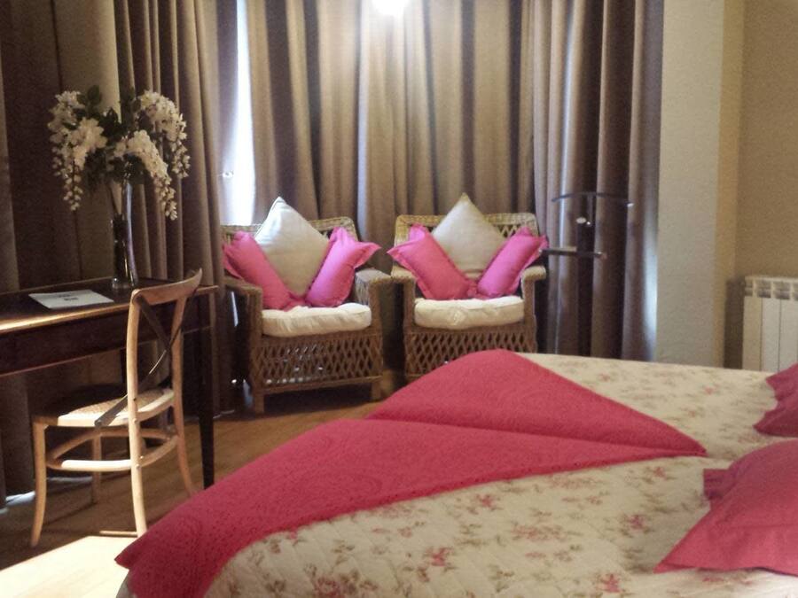 A hotel room with a bed, a desk, two wicker chairs with cushions, a vase with flowers, and a radiator.