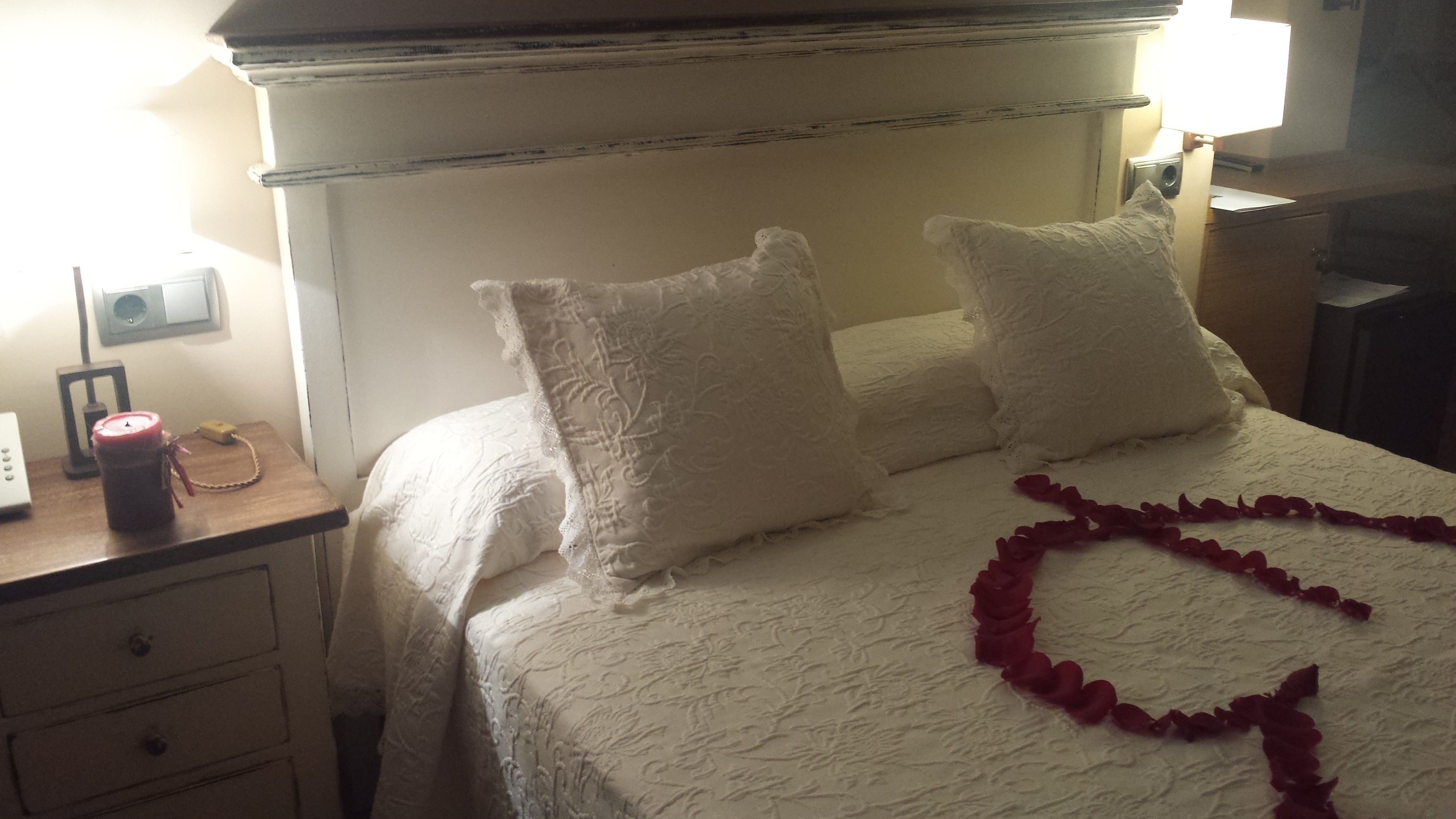 Una cama bien hecha con sábanas y almohadas blancas, una mesita de noche con una lámpara y una guirnalda decorativa de rosas sobre la cama.