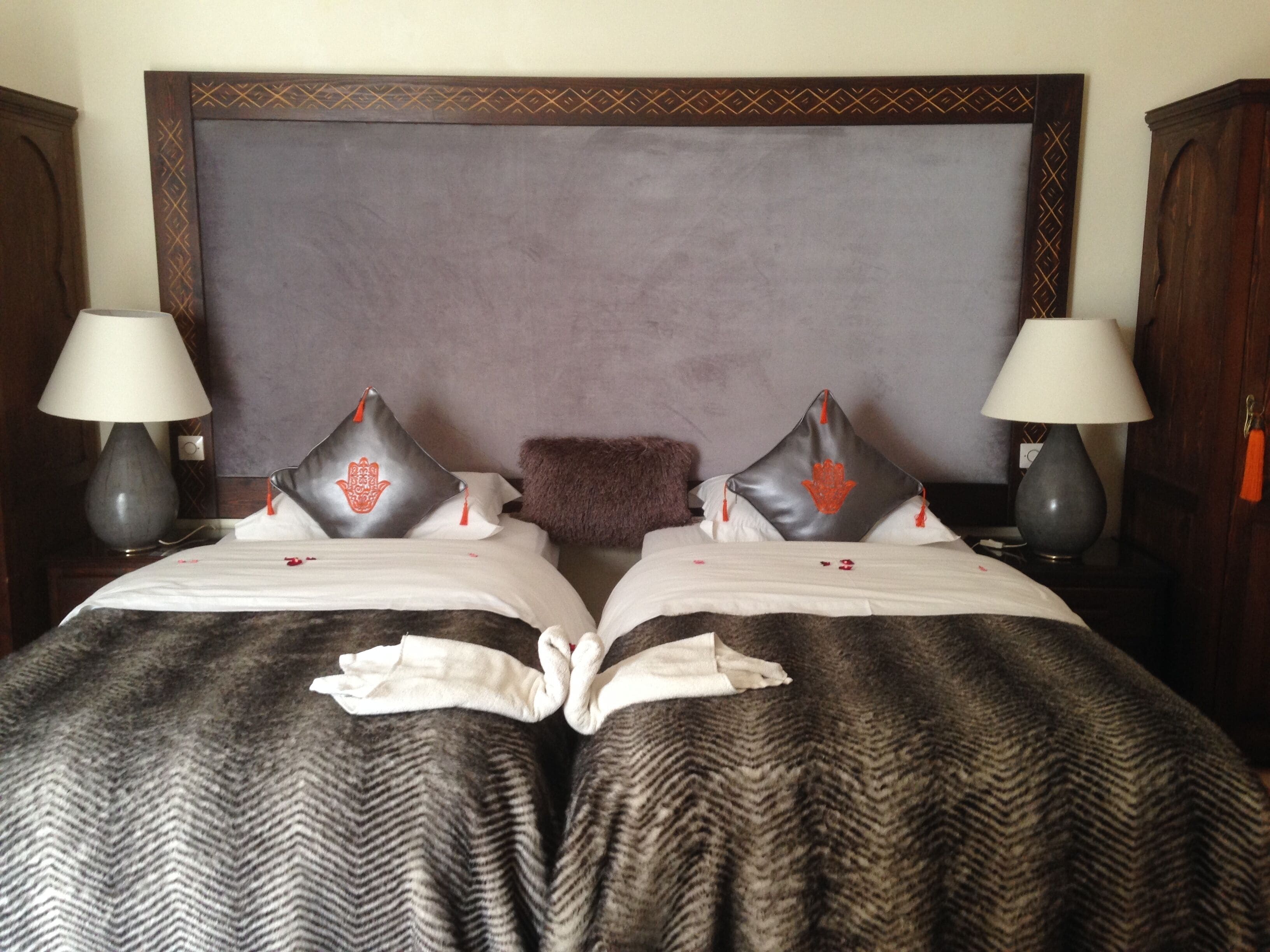 Un lit double avec une grande tête de lit sombre, deux lampes de chevet et des coussins décoratifs ornés d’un motif rouge.